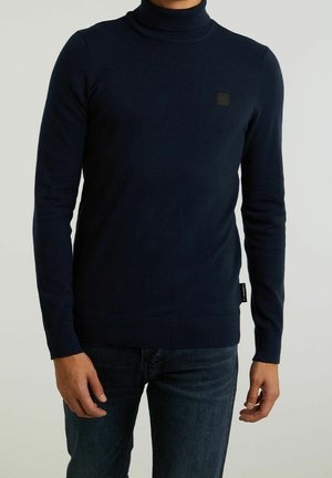 Man wearing a fitted dark navy turtleneck sweater and dark jeans, standing against a plain light background.