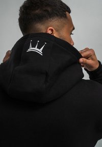 Der schwarze Kapuzenpullover hat ein Kapuze und ein gesticktes Kronenmotiv auf der Rückseite. Das Material wirkt weich mit einer glatten Textur.