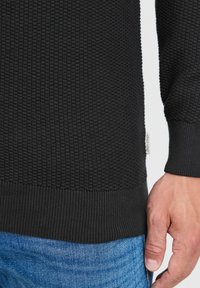 Svart teksturert genser med ribbestrikket kant. Synlig merkelapp på siden. Hånd som hviler nær livet med blå jeans delvis synlig.