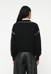 Pulôver preto de malha com uma bainha canelada e costura branca intrincada ao longo dos ombros. Combinado com calças de couro pretas. Design minimalista.