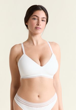 Donna con capelli scuri che indossa un reggiseno bianco senza cuciture e biancheria intima abbinata su uno sfondo neutro.