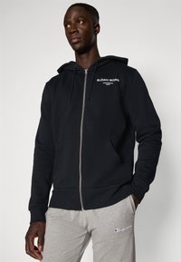 Schwarze Zip-Hoodie aus einem Baumwollmischgewebe mit einer Vorderseite, ausgestattet mit einem Fronttasche und weißem Logo-Text auf der linken Brustseite.