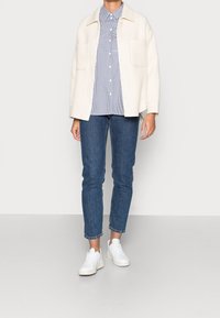 Krémová žebrovaná bunda s velkými kapsami, modro-bíle pruhovaná košile, úzké tmavé džíny a bílé tenisky.