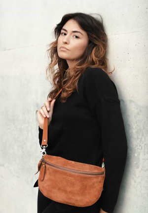 Junge Frau mit welligem Haar, die einen schwarzen Pullover trägt, eine braune Wildleder-Crossbody-Tasche trägt und sich an einer hellen Betonsohle lehnt.