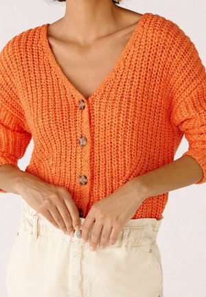 Persona che indossa un cardigan arancione testurizzato con bottoni e maniche arrotolate, che chiude il bottone di pantaloni panna a vita alta.