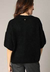 Pull en tricot noir avec des manches larges, finition texturée et un petit accent doré à l'arrière. Porté avec un pantalon gris à rayures.