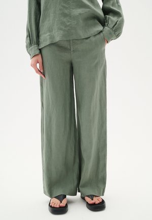 Pantalones de lino de pierna ancha en verde claro con un acabado texturizado, que cuentan con bolsillos laterales y un ajuste relajado; se combinan con sandalias planas negras.