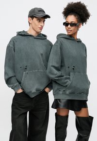 Zelená mikina s kapucňou v opotrebovanom štýle s predným vreckom, obsahuje veľké dierky; nosená dvomi modelkami, skombinovaná s tmavými džínsami, koženou sukňou a čižmami po kolená.