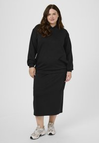 Femme grande taille portant un sweat à capuche noir et une jupe longue assortie avec des baskets beiges, se tenant contre un fond gris clair uni.