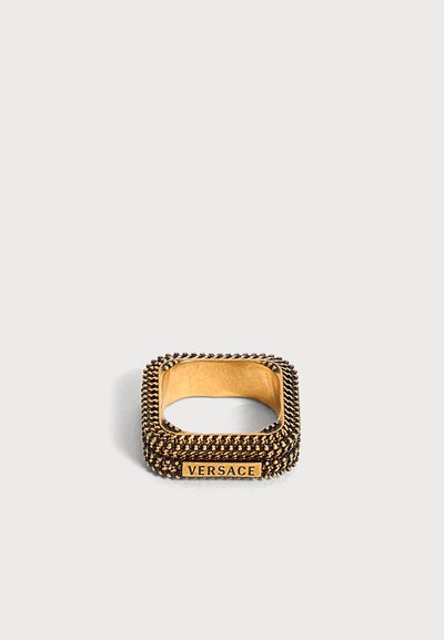 Τετράγωνο δαχτυλίδι Versace σε χρυσή απόχρωση με υφασμένους άκρους και φιρμάριστη πλάκα σε απλό ανοιχτό φόντο.