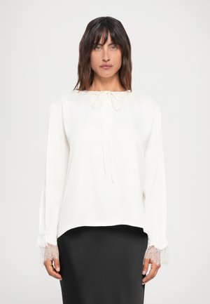 Bluză albă cu mâneci lungi, dantelă la manșete, croială lejeră, guler rotund cu detaliu de legare, textură netedă, asortată cu fustă neagră.