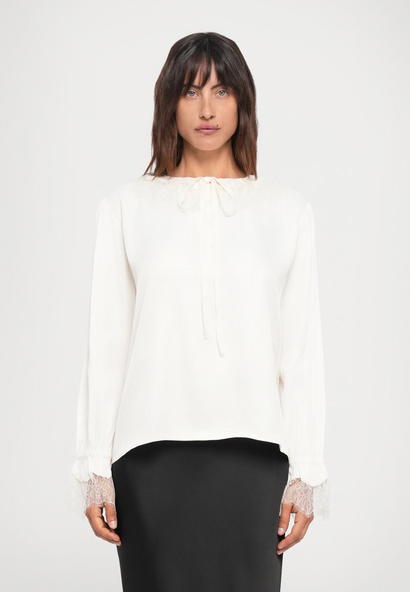 Weiße langärmlige Bluse mit Spitzenbesatz an den Ärmelbündchen, lockere Passform, runder Ausschnitt mit Bindung, glatte Textur, kombiniert mit einem schwarzen Rock.