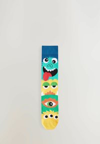 Chaussettes en coton colorées avec quatre visages de monstres en bande dessinée dans des tons de vert, jaune et orange, avec un bord côtelé bleu en haut.
