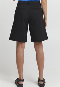 Svarta bomullsshorts med avslappnad passform, som har sidofickor och bakfickor samt en uppvikt fåll. Bärs med svarta sandaler, sedd bakifrån.