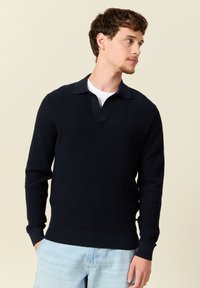 Navy neulepullover lyhyellä kauluksella sekä resoreilla hihoissa ja helmassa. Puvussa valkoinen t-paita, jossa on teksturoitu kangas.