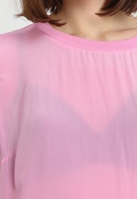 Jasnoróżowa, lekka bluzka o luźnym kroju i okrągłym dekolcie. Gładka tekstura z delikatnym gradientem koloru. Brak widocznych ozdób.