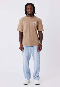 Camiseta marrón de manga corta con un logo frontal, combinada con unos vaqueros claros de corte holgado y zapatillas blancas. Fondo neutro.