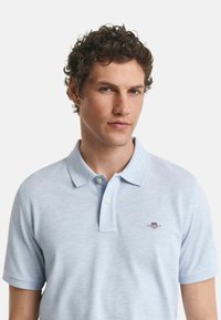 Man met krullend haar, gekleed in een lichtblauw polo-shirt met twee knopen en een klein geborduurd logo op de borst.