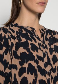 Blusa con motivo leopardato in beige e nero, caratterizzata da un collo alla mandarina, chiusura con bottoni e tessuto testurizzato. I punti salienti includono leggere pieghe.