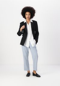 Črn blazer preko bele srajce s gumbom, v kombinaciji s svetlo modrimi hlačami. Črne loafersice dopolnjujejo videz. Minimalističen dizajn in struktura.