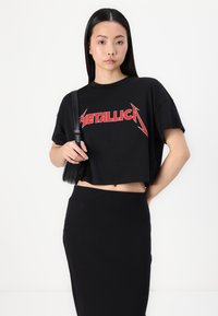 Črna skrajšana majica z rdečim napisom "METALLICA"; v kombinaciji s črno prilegajočo krilom. Model drži črno torbo z naramnico.