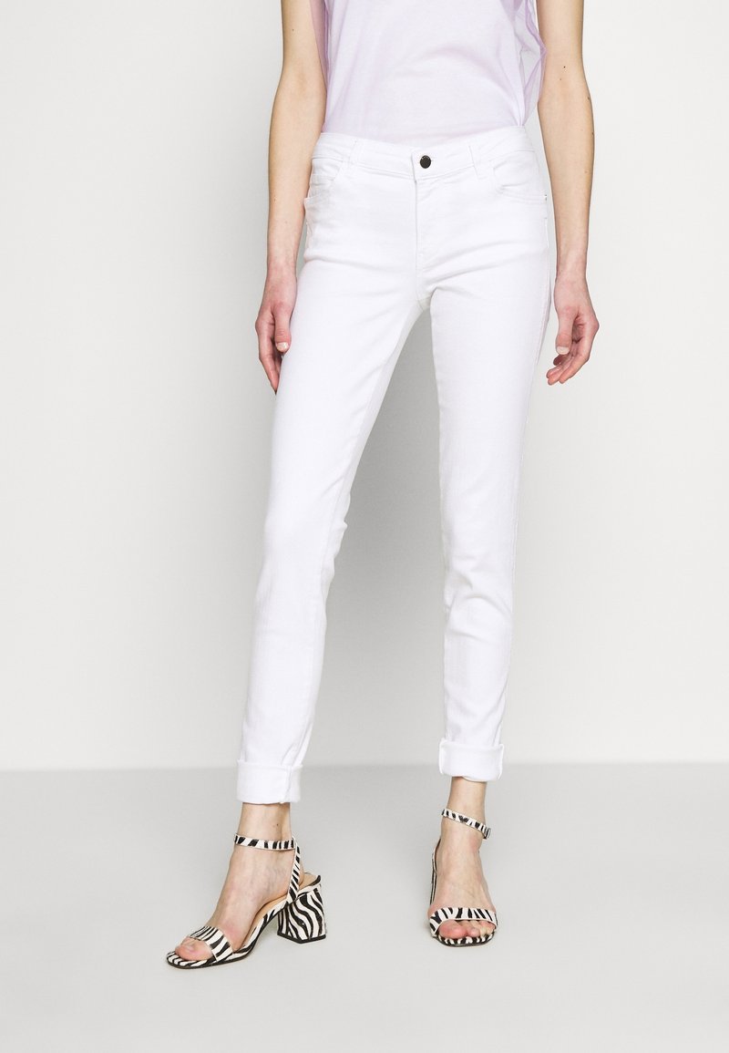Vaqueros de denim blanco con un corte ajustado, dobladillos acampanados y presillas para cinturón; combinados con sandalias de tacón de rayas en blanco y negro.