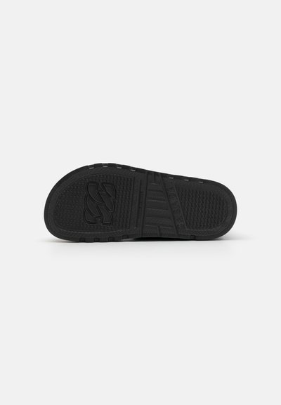 Semelle en caoutchouc noir avec adhérence texturée, présentant des stries et un logo proéminent. Conçue pour la durabilité avec un profil plat et une surface patinée.