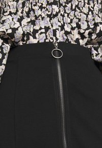 Jupe noire avec une fermeture éclair frontale et un tirette circulaire, associée à un chemisier noir à motifs floraux pastel avec un design froncé.