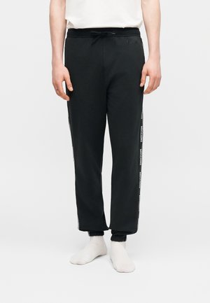 Mężczyzna noszący czarne dresy z elastycznymi ściągaczami i białymi paskami z logo po bokach, w połączeniu z białymi skarpetkami i białą koszulką.