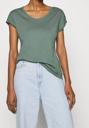 Henkilö pukeutuneena lyhythihaiseen vihreään t-paitaan, joka on kevyesti pistetty vaaleansinisiin korkeavyötäröisiin farkkuihin, seisoo yksiväristä taustaa vasten.