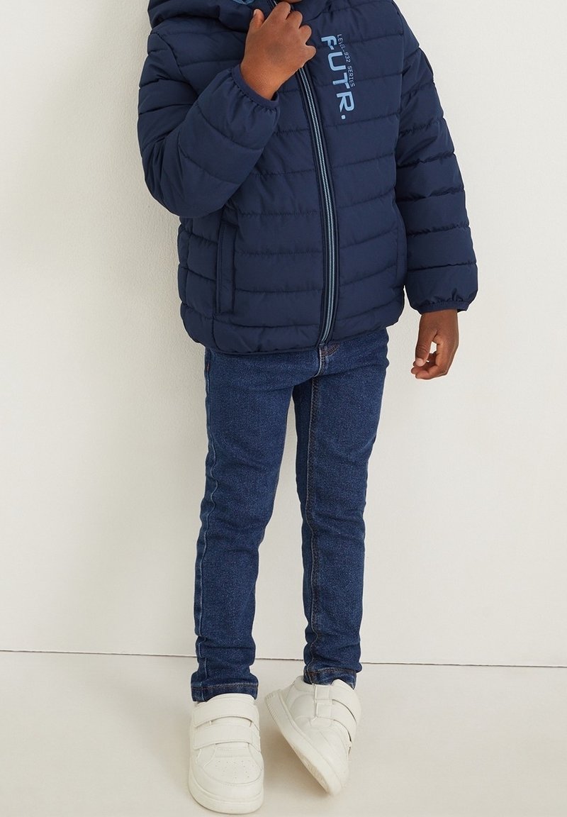 Enfant portant une doudoune bleu marine, un jean bleu et des baskets blanches à scratch, debout contre un mur clair.