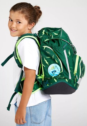 Smiling girl wearing white t-shirt and jeans carries large green backpack with yellow lightning patterns and cartoon character badge.