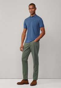 Modrá bavlněná polokošile, zelené chinos kalhoty a hnědé semišové boty. Polokošile má límečkový design a krátké rukávy. Kalhoty mají boční kapsy.