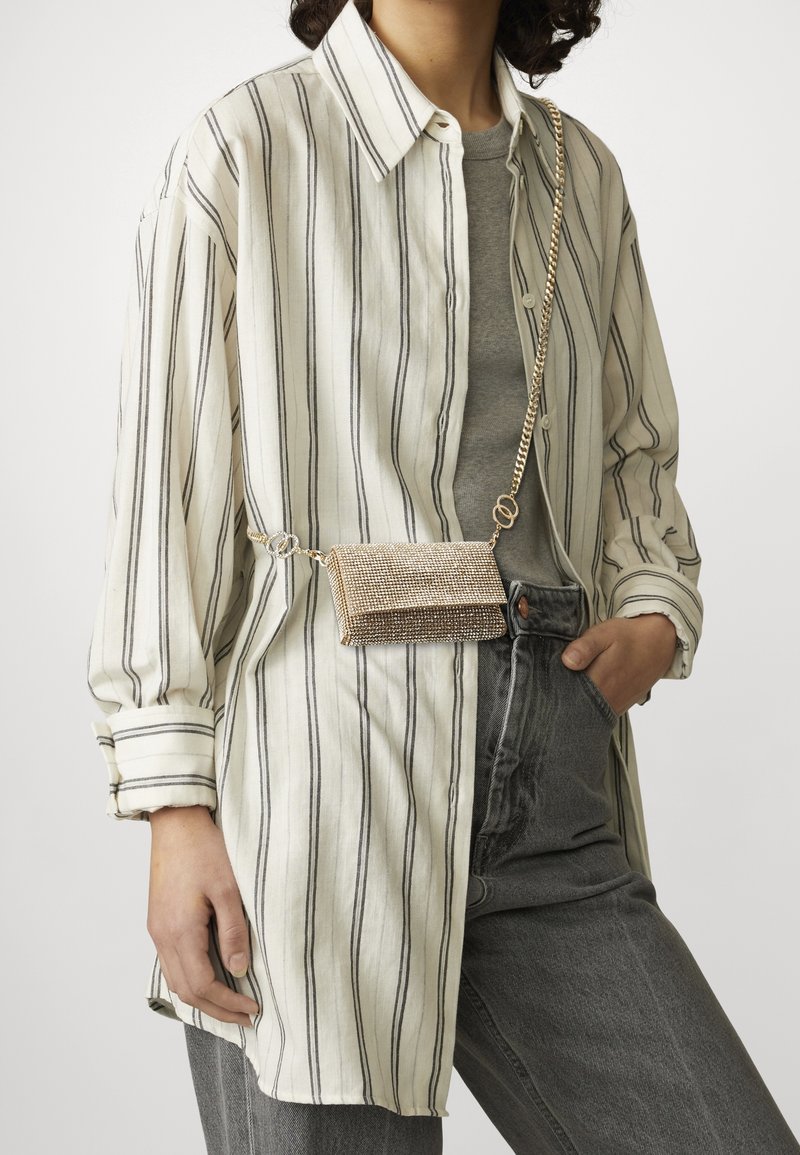 Metaliczna torba na ramię o teksturowanej złotej powierzchni, wyposażona w łańcuszkowy pasek i prostokątny kształt. Noszona na pasku nałożonym na pasiastą koszulę i szary top.