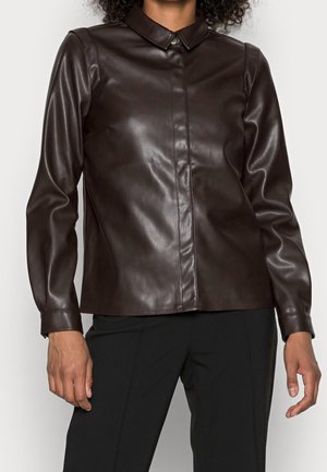Personne portant une chemise en cuir marron foncé à manches longues avec col, associée à un pantalon noir, debout devant un fond uni.