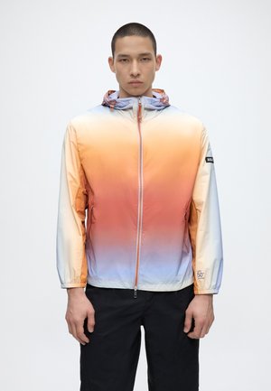 Vyras dėvi gradientinę ombre gobtuvu striukę oranžinės, rožinės ir šviesiai mėlynos spalvų deriniu, juodas kelnes, stovintis prieš paprastą baltą foną.