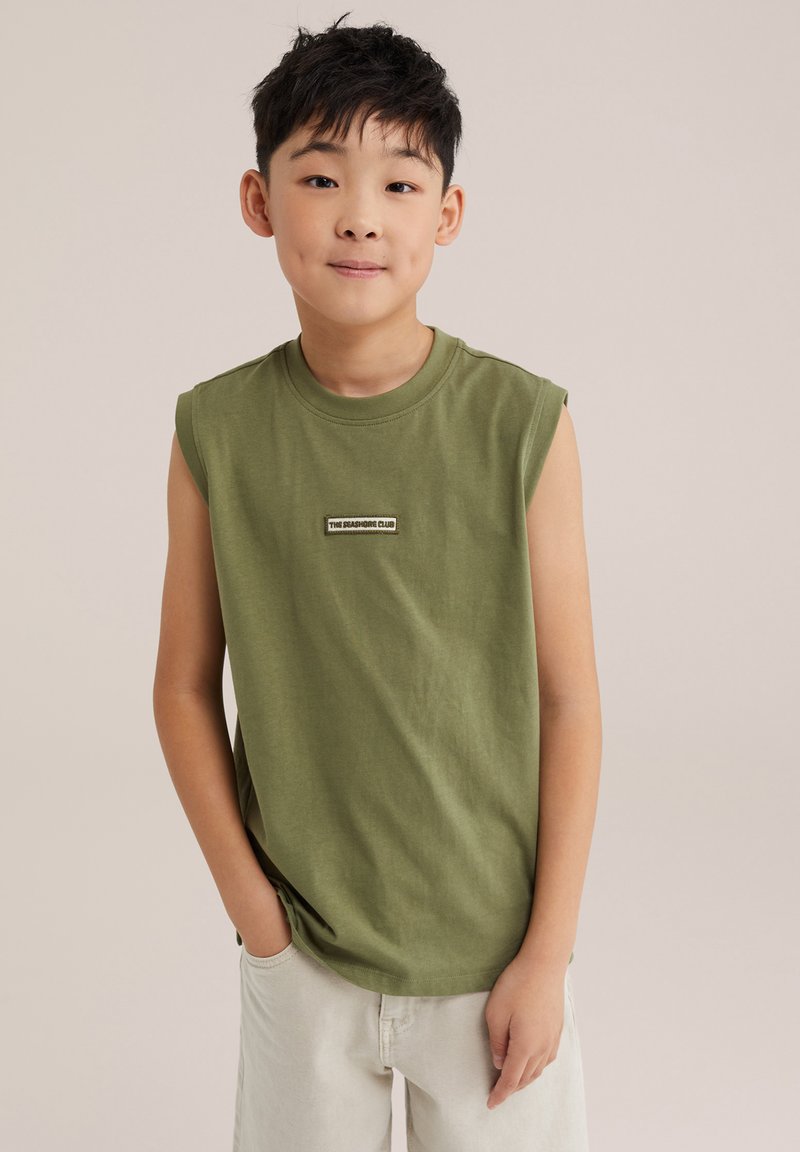 Garçon aux cheveux noirs courts portant une chemise sans manches vert olive et un pantalon beige, debout avec une main dans la poche devant un fond neutre.