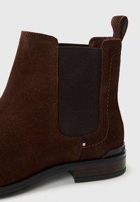 Bota Chelsea de ante marrón con panel lateral elástico negro, tirador en la parte posterior y tacón bajo de madera apilada con ribete negro en la suela.