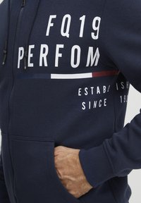 Sudadera con cremallera de color marino hecha de tela suave, que presenta un gran texto blanco 'FQ19 PERFORM' y acentos a rayas en rojo y azul. Incluye bolsillo tipo canguro.