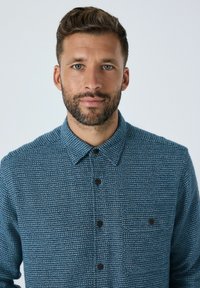 Mand med kort brunt hår og skæg iført en blå struktureret skjorte med knapper, vendt mod kameraet foran en ensfarvet lys baggrund.