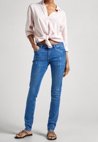 Camisa de botones de color rosa claro atada a la cintura, vaqueros ajustados azules con detalles desgastados y sandalias negras con acentos plateados.