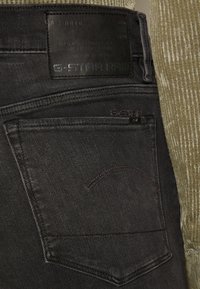 Jeans de mezclilla negra con un parche de cuero negro texturizado, que presenta el logo y detalles de cuidado; costuras visibles y un diseño de bolsillo sutil.