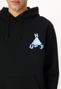 Black hoodie featuring a blue graphic design with "H," "U," and "F" letters and a triangular shape. Includes drawstrings and a front pocket.