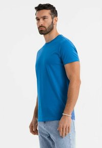 Modré tričko s krátkým rukávem vyrobené z hladké bavlny, vybavené kulatým výstřihem a přiléhavým střihem, doplněné světlými džíny.