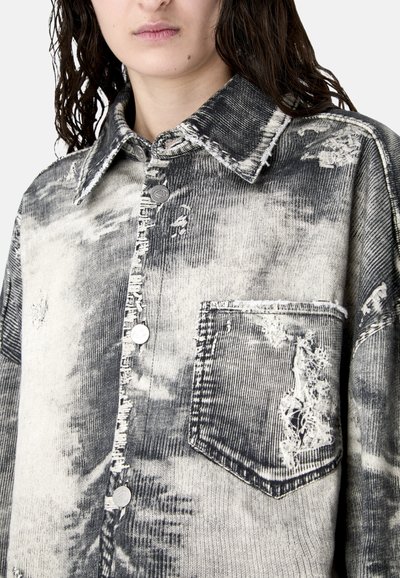 Frau trägt ein schwarz-weißes, acid-washed Cordhemd mit ausgefransten Details und einer großen Fronttasche.