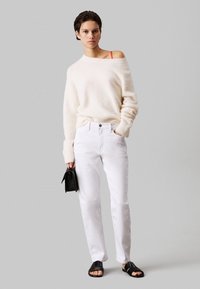 Kremowy, puszysty sweter z odkrytymi ramionami, białe proste dżinsy, czarne klapki, trzymając małą czarną torebkę.