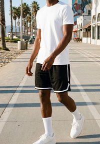 Homme marchant sur la promenade de la plage ensoleillée portant un t-shirt blanc, un short noir avec bordure blanche, des chaussettes blanches et des baskets blanches.