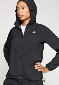 Veste noire légère avec capuche, dotée d’une fermeture éclair et de poignets élastiques. Comprend un logo réfléchissant sur la partie supérieure gauche de la poitrine.