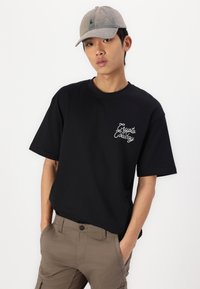 Camiseta negra de manga corta con logo en script blanco en el pecho, combinada con pantalones cargo marrón claro y una gorra gris desgastada.