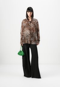 Mulher vestindo uma blusa de estampa de leopardo com calças largas pretas, segurando uma pequena bolsa verde, de pé contra um fundo branco.